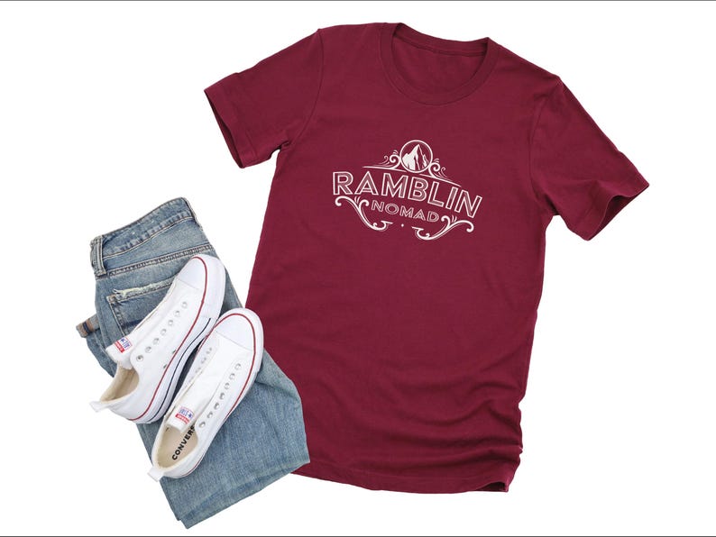 Puede incluir: Una camiseta color granate con texto blanco que dice "Ramblin Nomad". El texto est&aacute; en una fuente decorativa e incluye un peque&ntilde;o gr&aacute;fico de un pico de monta&ntilde;a.