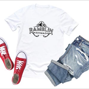 Puede incluir: Camiseta blanca con un dise&ntilde;o gr&aacute;fico negro que dice "Ramblin Nomad" con una monta&ntilde;a y un dise&ntilde;o floral. La camiseta se combina con jeans azules desgastados y zapatos de lona rojos.