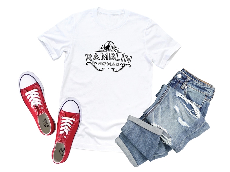Puede incluir: Camiseta blanca con un dise&ntilde;o gr&aacute;fico negro que dice "Ramblin Nomad" con una monta&ntilde;a y un dise&ntilde;o floral. La camiseta se combina con jeans azules desgastados y zapatos de lona rojos.