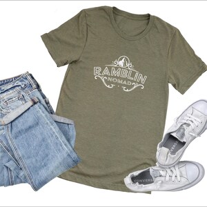 Puede incluir: Una camiseta verde con el texto "Ramblin Nomad" en letras blancas. La camiseta se combina con unos vaqueros azules y unas zapatillas blancas.