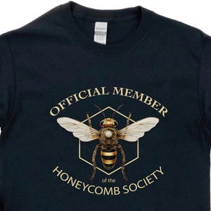 May include: Navy blue long-sleeve shirt featuring a detailed illustration of a bee within a gold hexagonal frame. The text reads "OFFICIAL MEMBER of the HONEYCOMB SOCIETY" in gold lettering. The shirt is made of a soft material.