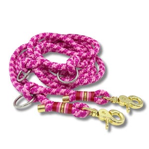 May include: A pink and fuchsia rope dog lead with gold-coloured clasps and metal rings. The lead is made of thick, braided rope and features decorative bands of red, pink, and gold near the clasps. The lead is designed for pet owners.