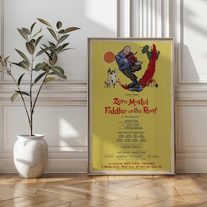 May include: A framed vintage poster for the musical "Fiddler on the Roof" featuring Zero Mostel. The poster has a yellow background with illustrations of a fiddler, a dog, and a red streak. The text includes the musical's title and the names of the creators.