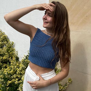 May include: A blue crochet crop top with a textured pattern. The top is sleeveless and paired with white drawstring pants. The outfit is worn outdoors, with a background of greenery and a neutral-toned wall.