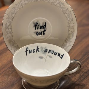 May include: A white porcelain teacup and saucer set featuring a delicate grey floral pattern and silver trim. The teacup interior displays the black handwritten text 'fuck around', while the saucer center features the text 'find out'.