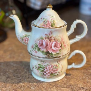 May include: A cream-colored teacup and teapot set. The set features pink roses and green leaves painted on the exterior, with gold trim. The teapot sits atop the teacup, creating a stacked design.