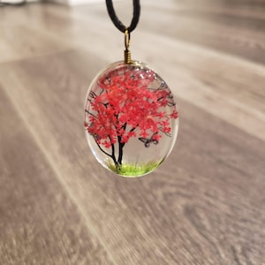 May include: A clear oval pendant necklace with a red flower tree and two butterflies inside. The pendant is on a black cord.