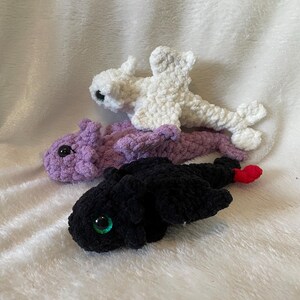 May include: Three crocheted dragon plushies. One is white, one is purple, and one is black with green eyes.