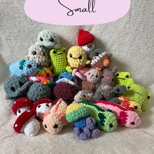 May include: A collection of small, handmade crochet plush toys in various shapes and colors, including frogs, turtles, mushrooms, and axolotls. The word "Small" is written in cursive on a pink oval.