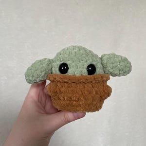 Peut inclure: Une peluche faite &agrave; la main repr&eacute;sentant un personnage avec de grands yeux noirs, de grandes oreilles et un v&ecirc;tement marron textur&eacute;. Le jouet est principalement vert clair et marron, avec une texture douce et floue. Le jouet est tenu dans une main.