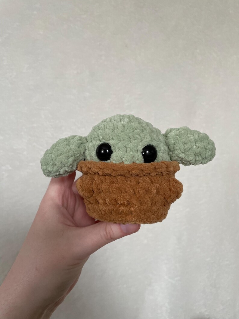 Peut inclure: Une peluche faite &agrave; la main repr&eacute;sentant un personnage avec de grands yeux noirs, de grandes oreilles et un v&ecirc;tement marron textur&eacute;. Le jouet est principalement vert clair et marron, avec une texture douce et floue. Le jouet est tenu dans une main.