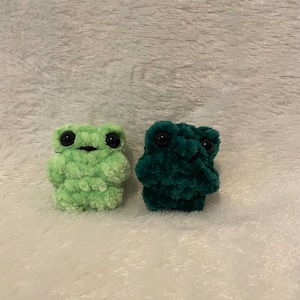 May include: Two small crocheted frog plushies, one green and one a darker shade of green. Both have black eyes.