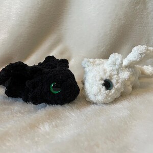 May include: Two crocheted plush toys shaped like dragons. One is black with green eyes and the other is white with black eyes.