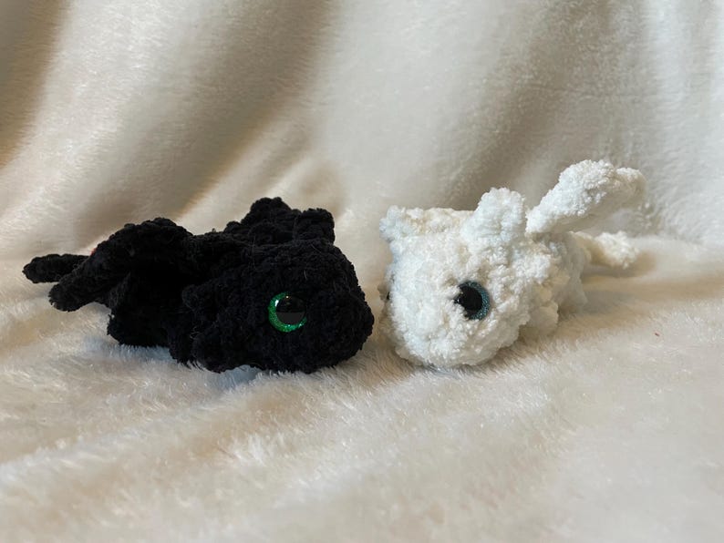 May include: Two crocheted plush toys shaped like dragons. One is black with green eyes and the other is white with black eyes.
