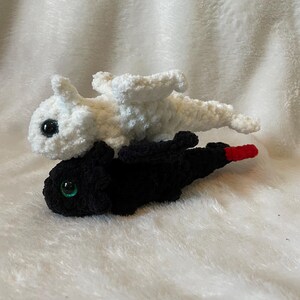 May include: Two crocheted plush dragons, one white and one black, with green eyes. The white dragon is on top of the black dragon.