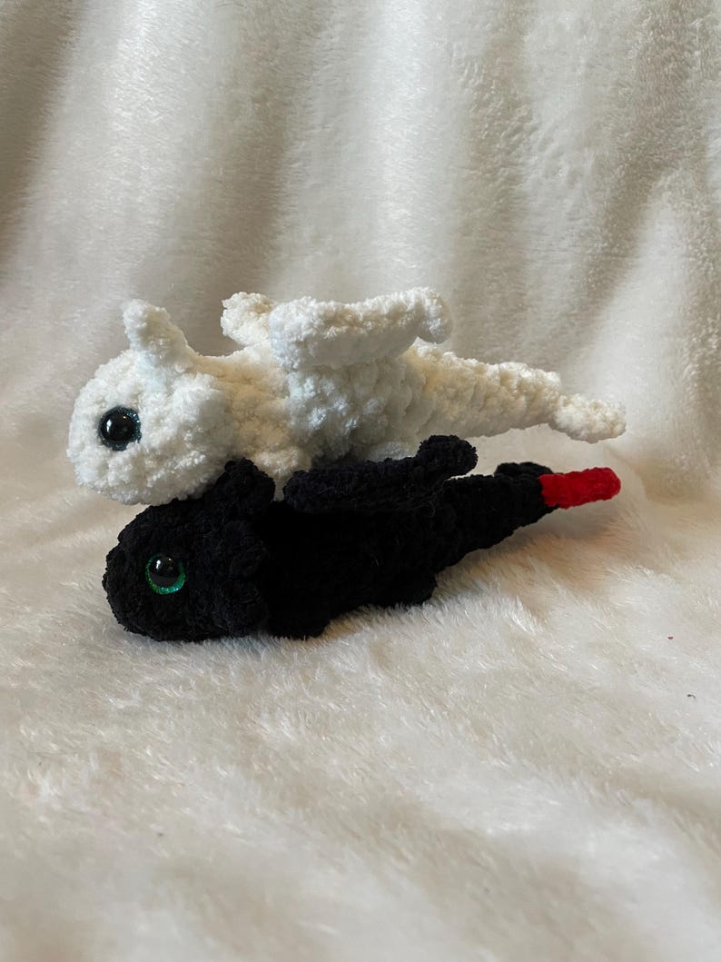May include: Two crocheted plush dragons, one white and one black, with green eyes. The white dragon is on top of the black dragon.