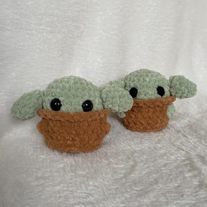 Peut inclure: Deux figurines faites &agrave; la main au crochet repr&eacute;sentant un personnage avec de grandes oreilles et des yeux noirs. Les figurines sont vert clair avec des v&ecirc;tements marron. Les figurines sont pos&eacute;es sur une surface blanche textur&eacute;e.