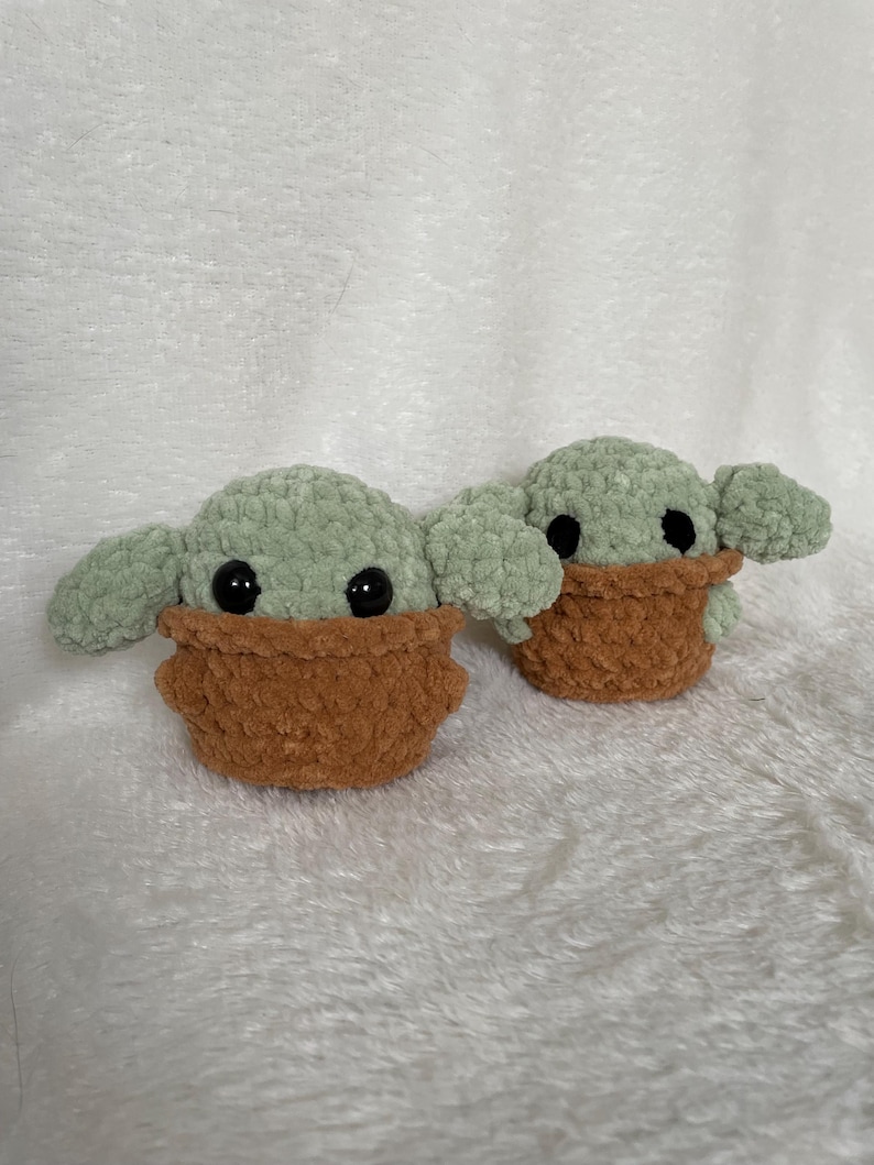 Peut inclure: Deux figurines faites &agrave; la main au crochet repr&eacute;sentant un personnage avec de grandes oreilles et des yeux noirs. Les figurines sont vert clair avec des v&ecirc;tements marron. Les figurines sont pos&eacute;es sur une surface blanche textur&eacute;e.
