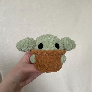Peut inclure: Une peluche faite &agrave; la main repr&eacute;sentant un personnage avec de grandes oreilles et des yeux noirs, dans un porte-b&eacute;b&eacute; marron. Le jouet est fait de fil doux et textur&eacute; dans les tons de vert et de marron.