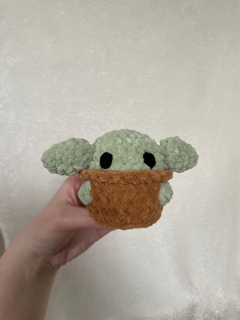 Peut inclure: Une peluche faite &agrave; la main repr&eacute;sentant un personnage avec de grandes oreilles et des yeux noirs, dans un porte-b&eacute;b&eacute; marron. Le jouet est fait de fil doux et textur&eacute; dans les tons de vert et de marron.