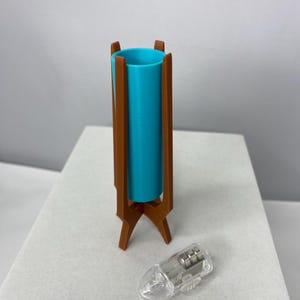 May include: A teal cylinder lamp with a brown wooden stand. The lamp has a clear plastic light bulb next to it. The lamp is a mid-century modern design.