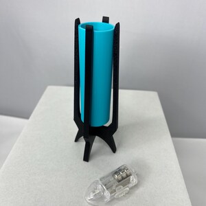 May include: A teal cylinder vase in a black, three-legged stand. A clear, teardrop-shaped light bulb is next to the vase. The vase is approximately 6 inches tall.