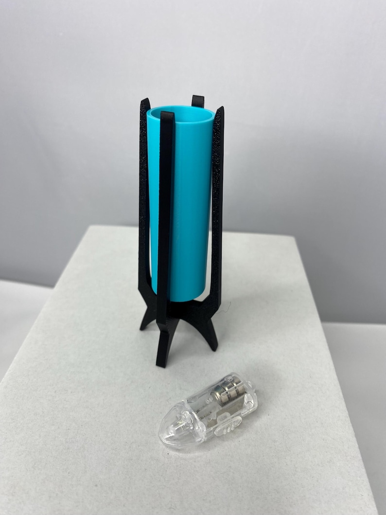 May include: A teal cylinder vase in a black, three-legged stand. A clear, teardrop-shaped light bulb is next to the vase. The vase is approximately 6 inches tall.