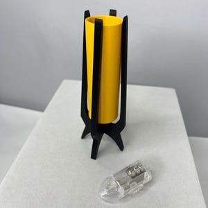 May include: A yellow cylindrical lamp with a black, three-legged base and a clear, teardrop-shaped light bulb. The lamp has a modern, minimalist design. The lamp is approximately 6 inches tall.