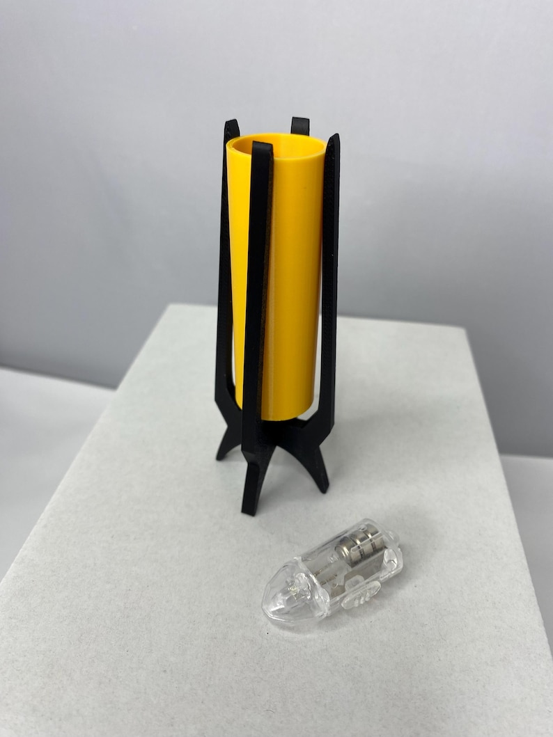 May include: A yellow cylindrical lamp with a black, three-legged base and a clear, teardrop-shaped light bulb. The lamp has a modern, minimalist design. The lamp is approximately 6 inches tall.