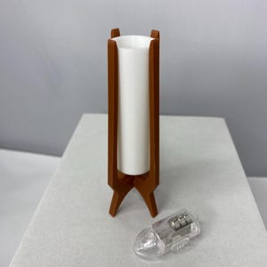 May include: A mid-century modern style lamp with a brown wooden frame and a white cylindrical shade. The lamp has a tripod base. A small, clear plastic object is next to the lamp.