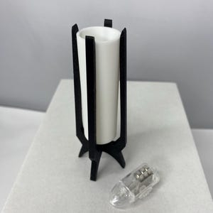 May include: A white cylindrical candle holder with a black, geometric stand. The stand has three legs and vertical supports. A small, clear light bulb is next to the candle holder. The candle holder is approximately 4 inches tall.