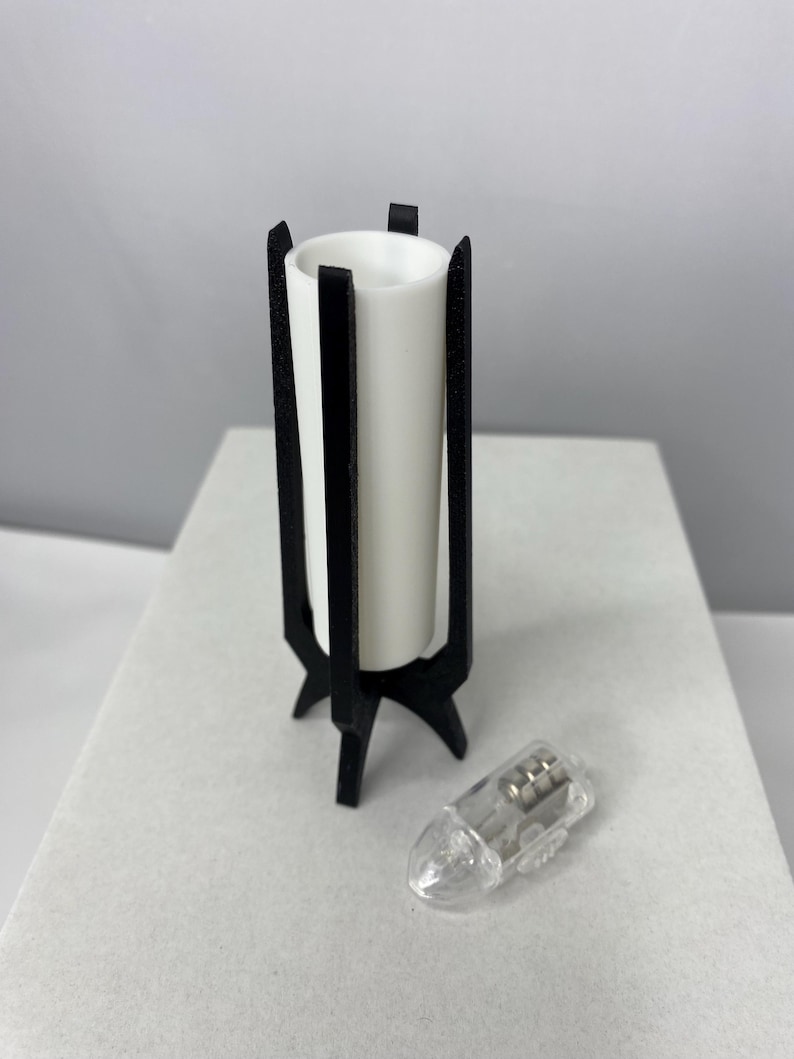 May include: A white cylindrical candle holder with a black, geometric stand. The stand has three legs and vertical supports. A small, clear light bulb is next to the candle holder. The candle holder is approximately 4 inches tall.