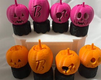 May include: A collection of eight 3D-printed pumpkins in bright pink and orange, each resting on a black cylindrical base. Four pumpkins feature carved faces, while others display a letter B or a silhouette figure. They are arranged in two rows.