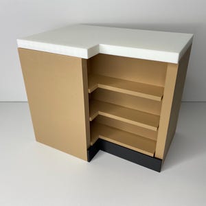 May include: A tan-colored, corner display cabinet with a white countertop. The cabinet has three shelves and a black base. The design is modern and minimalist, suitable for retail or home use.