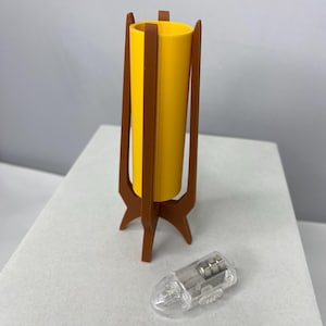 May include: A miniature yellow lamp with a brown wooden stand and a clear lightbulb. The lamp has a cylindrical shade and a tripod base. The lightbulb is clear and has a small metal component inside. The lamp is designed in a mid-century modern style.