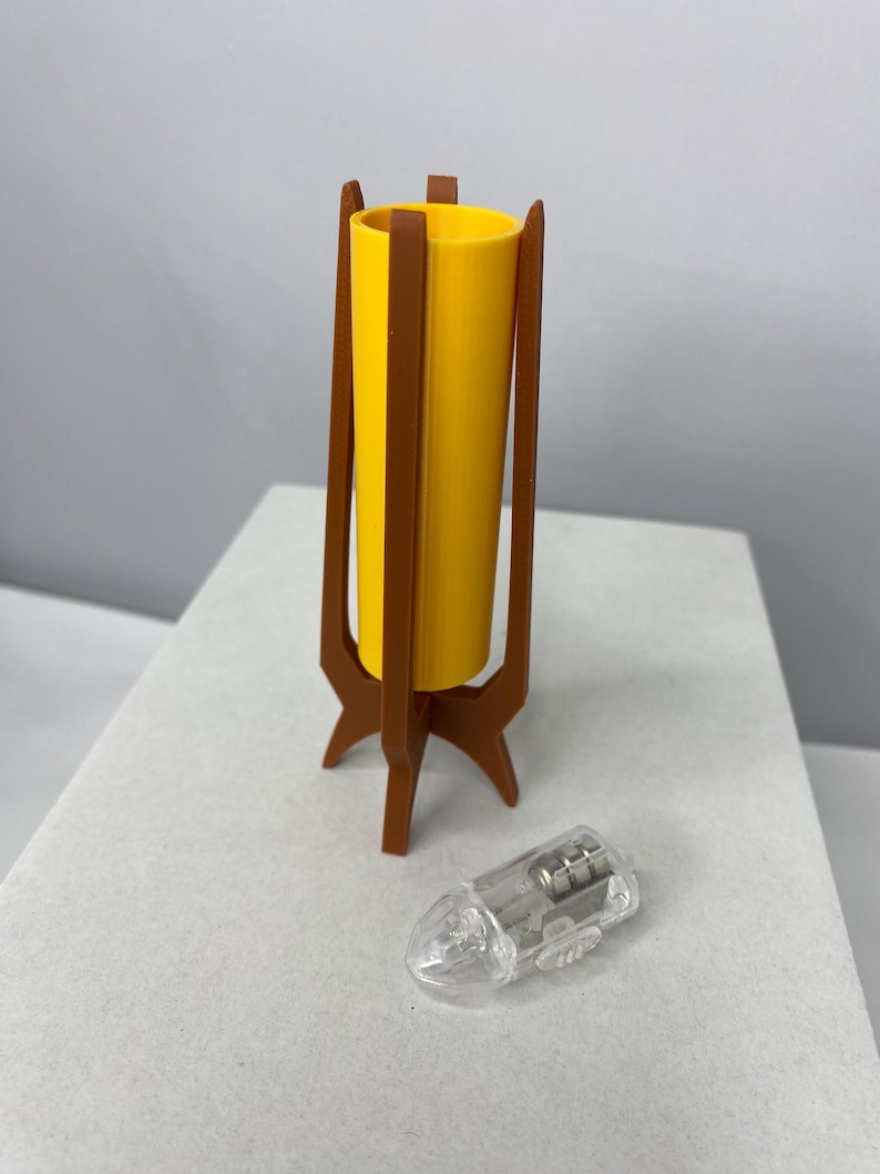 May include: A miniature yellow lamp with a brown wooden stand and a clear lightbulb. The lamp has a cylindrical shade and a tripod base. The lightbulb is clear and has a small metal component inside. The lamp is designed in a mid-century modern style.