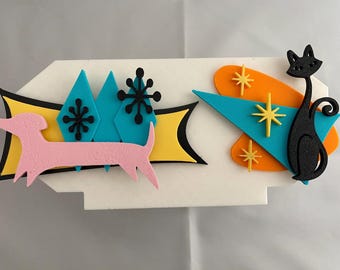 May include: A white rectangular wall plaque featuring a pink dachshund silhouette, a black cat silhouette, and colorful geometric shapes including blue diamonds, yellow starbursts, and orange ovals arranged in a layered, multi-dimensional design.