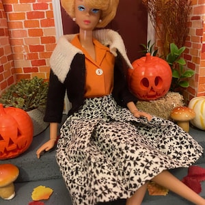 May include: A blonde doll wearing a black jacket with a white collar, an orange blouse, and a black and white floral skirt. The doll is sitting on a set of steps with a carved pumpkin and fall leaves around her.