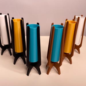 May include: A collection of mid-century modern style vases. The vases feature a cylindrical design with a variety of colors, including white, yellow, and teal. The holders are black and brown, with a tripod base.