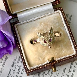 May include: A gold-toned ring featuring a fox head with red gemstone eyes. The ring is displayed in a vintage brown jewelry box with a cream-colored interior. A purple flower is visible in the background.