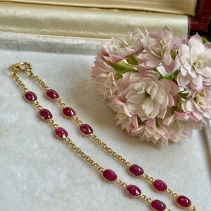 Może przedstawiać: Złota bransoletka z owalnymi, głęboko czerwonymi kamieniami szlachetnymi. Bransoletka jest prezentowana na białej powierzchni obok bukietu różowych kwiatów. Zapięcie jest widoczne, a kamienie są równomiernie rozmieszczone na łańcuszku.