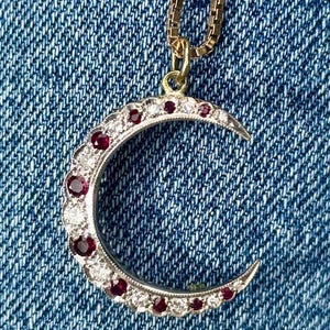 Op de afbeelding: Een halve maan hanger aan een goudkleurige ketting. Het sieraad heeft een zilverkleurig metalen frame dat is bezet met afwisselend ronde rode robijnen en heldere witte diamanten die langs de gebogen vorm van de maan zijn geplaatst.