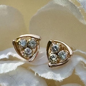 May include: A pair of gold stud earrings, each featuring a triangular frame that encloses three small, round, clear faceted stones arranged in a cluster. The metal has a warm, polished finish, and the stones exhibit a bright, reflective sparkle.