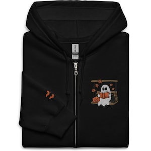 Peut inclure: Sweat-shirt à capuche noir avec fermeture éclair, orné d'un motif brodé représentant un fantôme lisant un livre devant une cheminée. Le fantôme porte un chapeau de citrouille et il y a des chauves-souris et des feuilles qui tombent autour de lui.