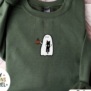 May include: A military green sweatshirt with an embroidered design of a ghost holding a mug and a black cat. The text "Cozy Seasons Apparel Est 2023" is on the bottom left corner of the sweatshirt. The text "Embroidered" is on the bottom right corner of the sweatshirt. The text "Military Green" is on the top right corner of the sweatshirt.