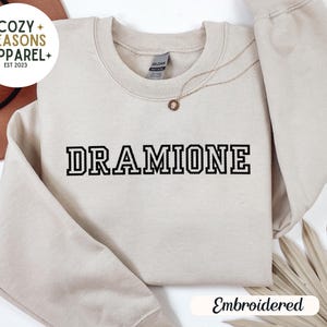 May include: Sand coloured sweatshirt with the text "DRAMIONE" embroidered in black. The sweatshirt is a crew neck style and has a relaxed fit. The text is in a bold, blocky font.
