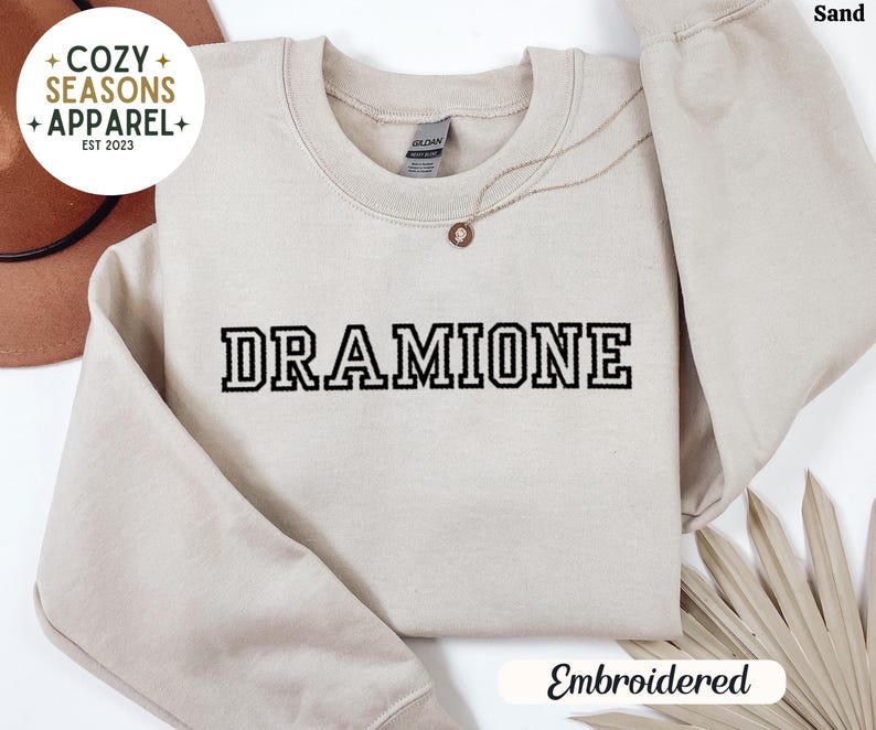 May include: Sand coloured sweatshirt with the text "DRAMIONE" embroidered in black. The sweatshirt is a crew neck style and has a relaxed fit. The text is in a bold, blocky font.