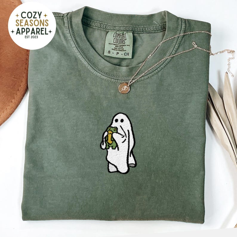 May include: A green t-shirt with a white ghost holding a green frog embroidered on the front. The shirt has a tag that says "COMFORT COLORS" and "S - P - CH".