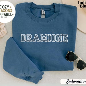 May include: Indigo blue sweatshirt with the word "DRAMIONE" embroidered in white on the front. The sweatshirt is folded and laid flat on a white surface. The word "Embroidered" is written in black text below the sweatshirt.