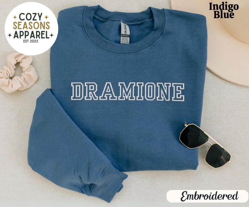 May include: Indigo blue sweatshirt with the word "DRAMIONE" embroidered in white on the front. The sweatshirt is folded and laid flat on a white surface. The word "Embroidered" is written in black text below the sweatshirt.